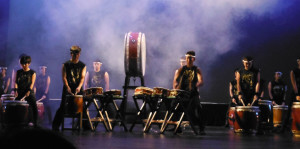 Large taiko drum on stage with students at smaller drums and illuminated theater fog