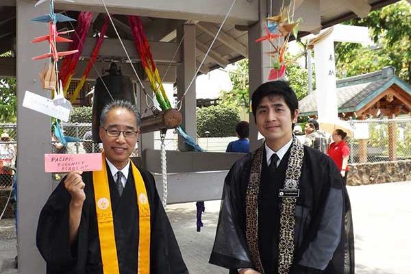 Bishop Eric Matsumoto and Hawaii Betsuin minister Rev. Joshin Kamuro at Hiroshima Peace Bell with strings of cranes with attached sign saying "Pacific Buddhist Academy"