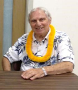Rev. Dr. Alfred Bloom, seated, smiling, wearing yellow-orange lei