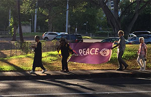 peace walk participants walk on Punchbowl St. side walk with purple PEACE banner backlit by evening sun