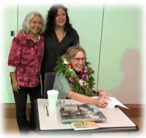 Pat Masters, smiling and wearing lei, signing a book with two women friends standing behind