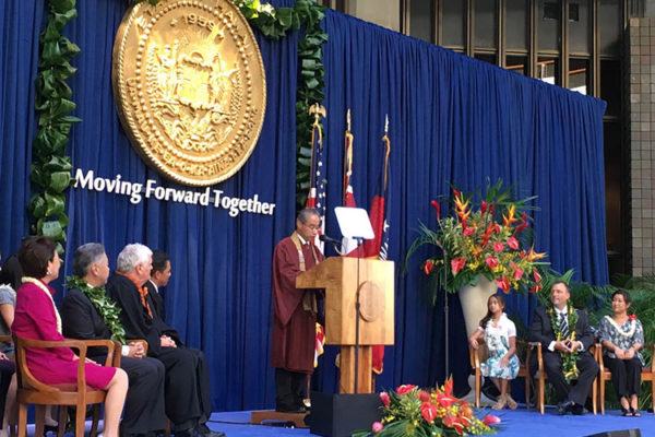 Bishop Matsumoto at podium on stage with Gov. David Ige and Lt. Gov. Josh Green and others seated on stage