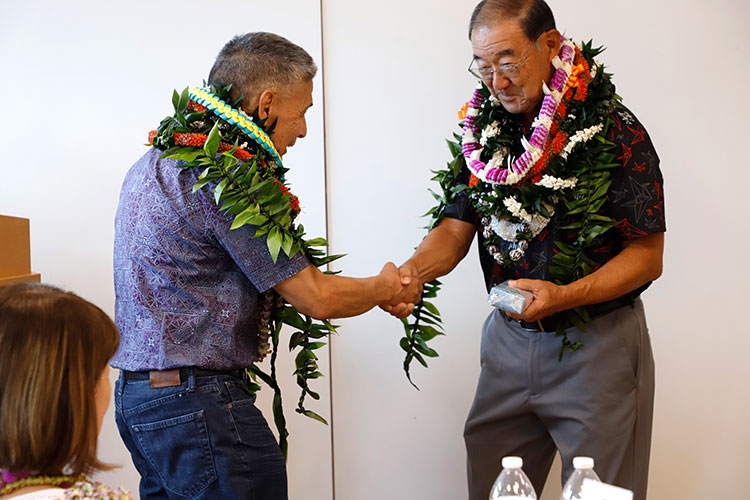 two men wearing many lei shake hands and slightly bow
